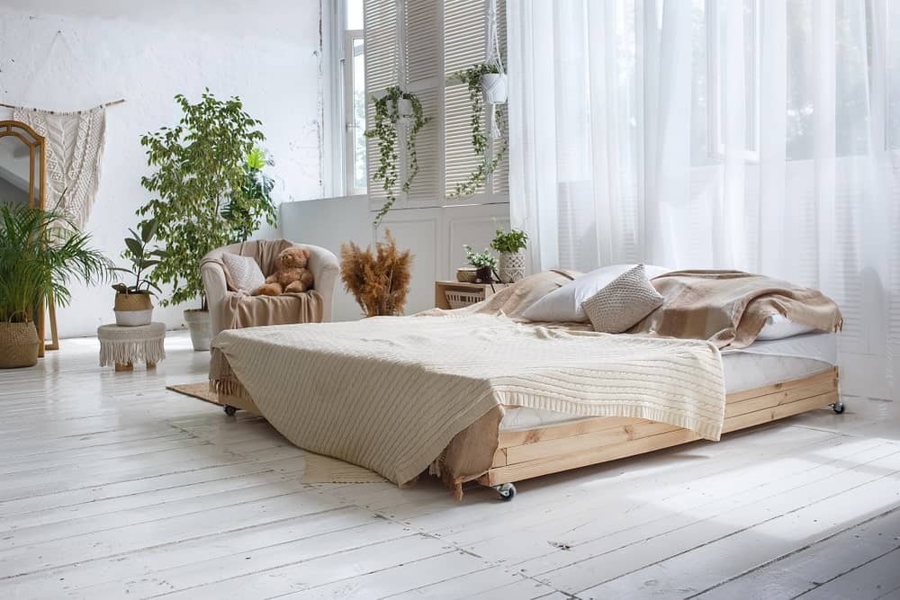 Light and airy bedroom offers a wooden bed with wheels accompanied by a matching nightstand and white round back chair. It has distressed wide plank flooring and glazed windows covered in flowy sheer curtains.