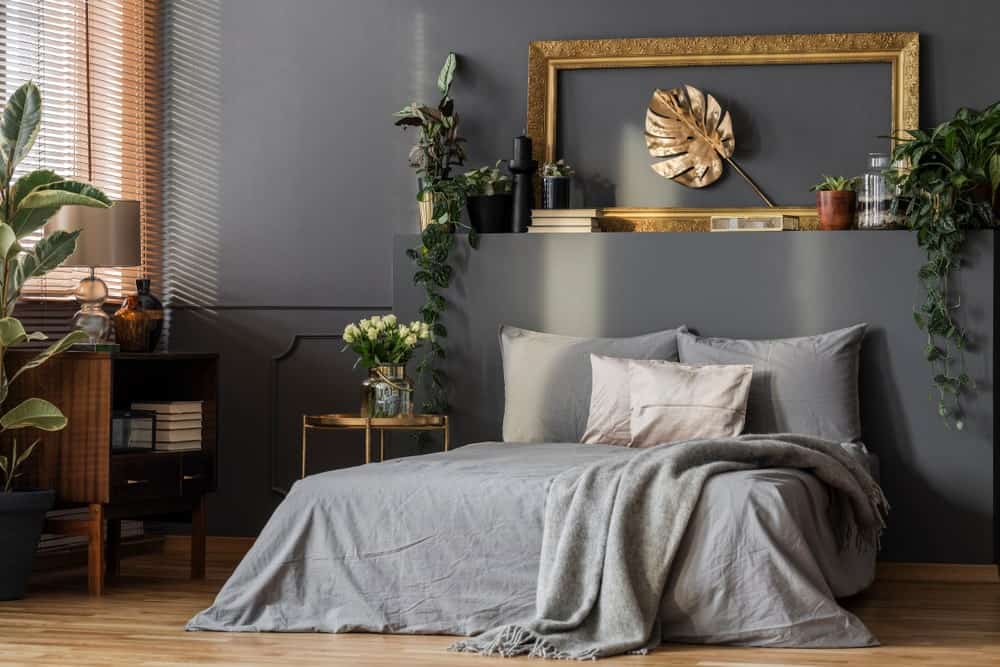 This primary bedroom is decorated with potted plants and a lovely leaf wall art framed in ornate gold. It is mounted above the platform bed that's dressed in gray bedding.