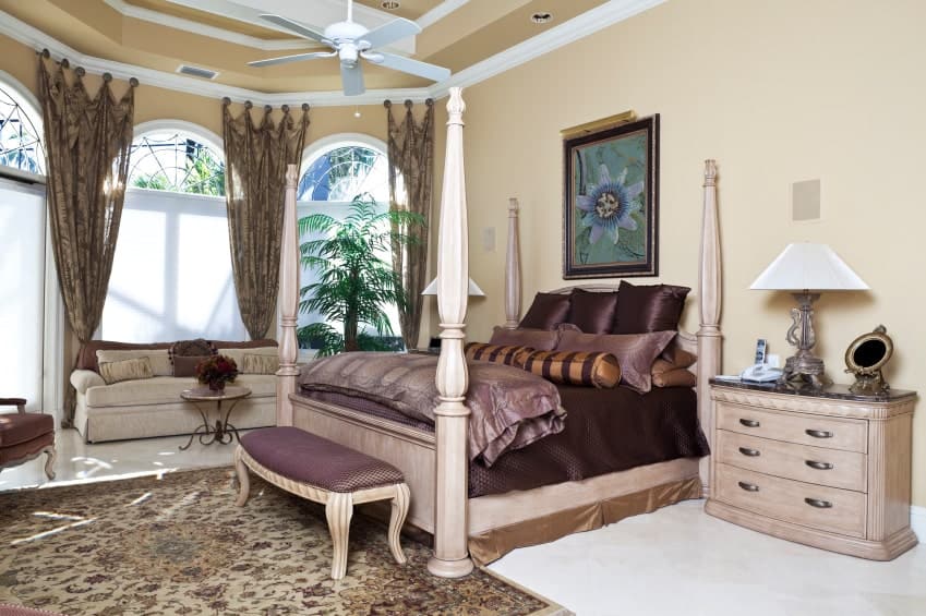 Elegant primary bedroom showcases a wooden four poster bed and a beige skirted sofa by the arched windows dressed in tropical curtains. It includes floral wall art and a white fan that hung from the tray ceiling.