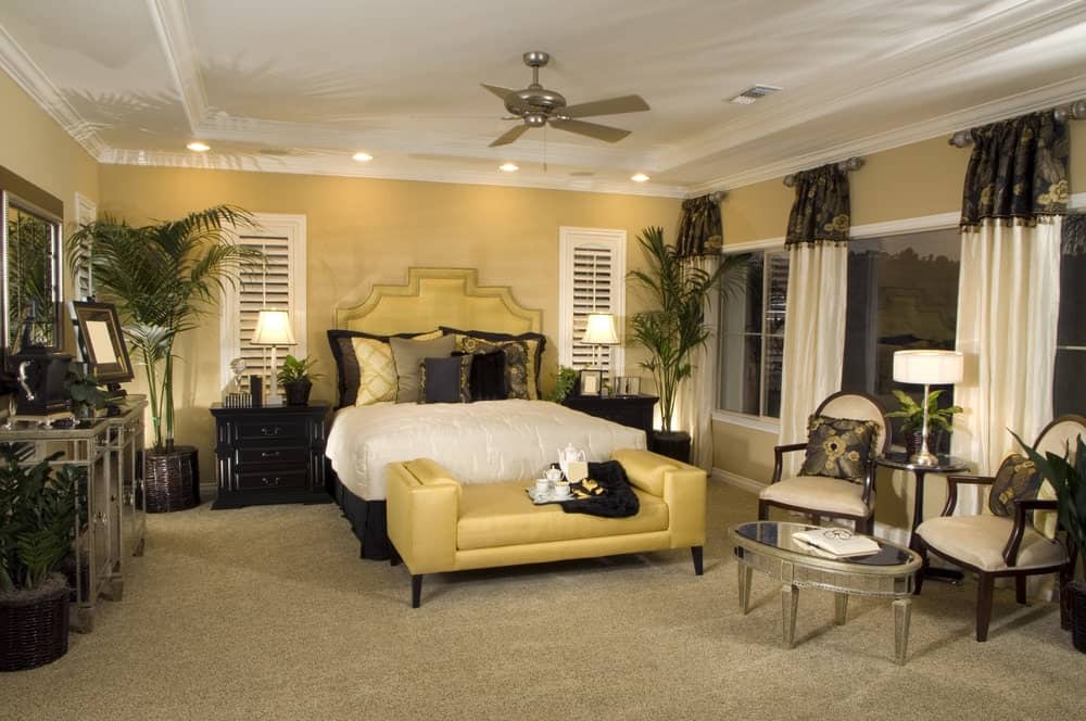 A yellow upholstered bench sits in front of the skirted bed in this primary bedroom with a seating area showcasing cushioned chairs and an oval coffee table.