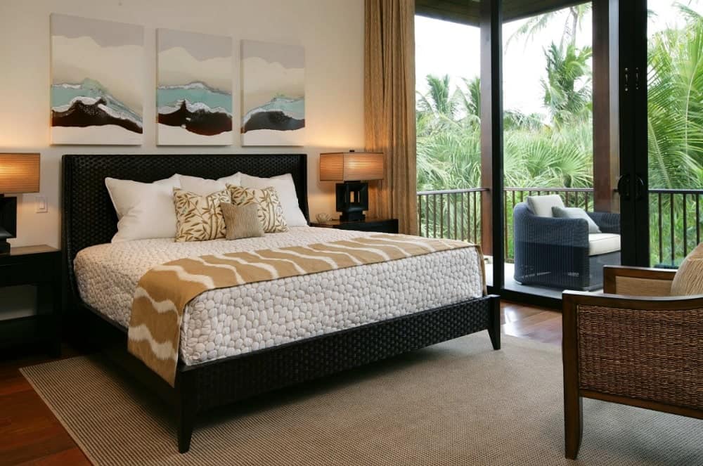 Lovely wall arts hang above the black textured bed dressed in pebble bedding in this primary bedroom with sleek table lamps and a wicker armchair that sits on a tan rug.