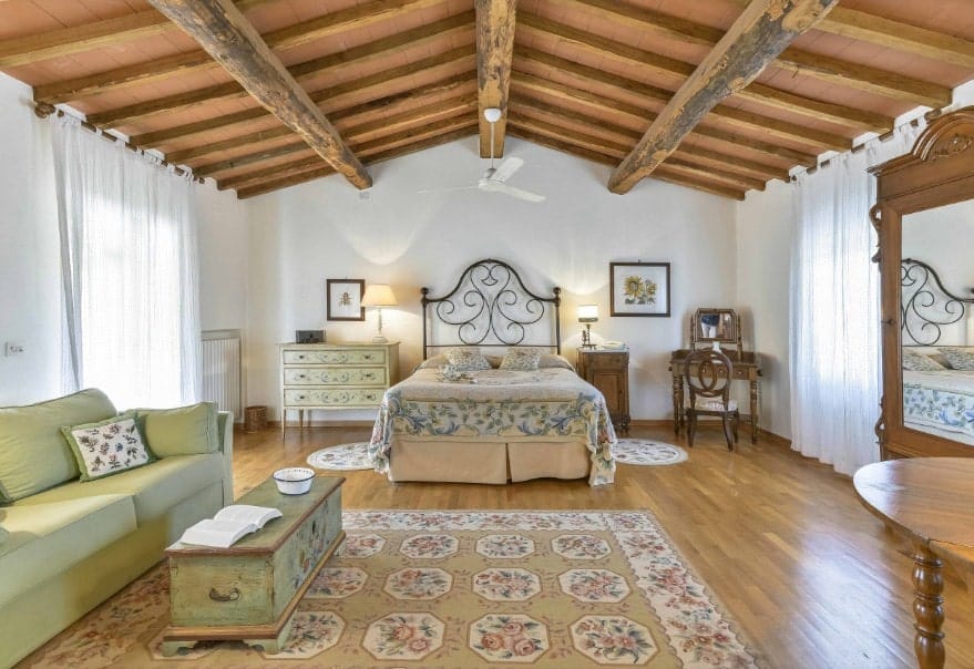 Spacious primary bedroom featuring a wooden ceiling with exposed beams along with hardwood flooring. The room offers a classy bed along with an olive green couch on the side.