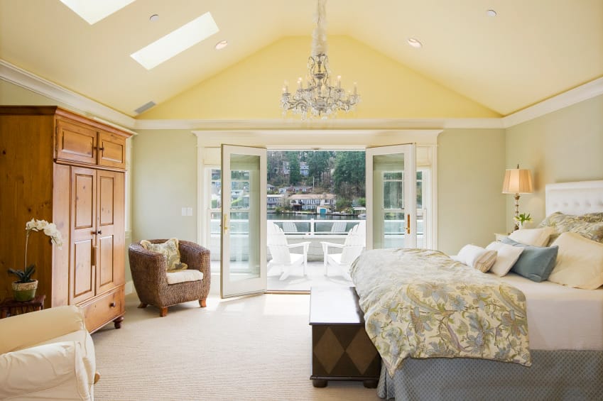 Spacious primary bedroom featuring a vaulted ceiling with skylights and carpet flooring. The room has a large elegant bed lighted by table lamps and a gorgeous chandelier.
