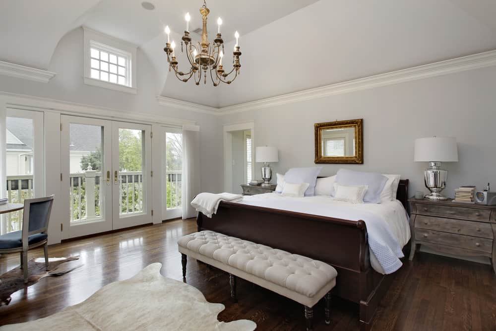 Large primary bedroom featuring light gray walls along with hardwood flooring. The room has a large bed with rustic bedside tables on both sides. The room is lighted by a gorgeous chandelier.
