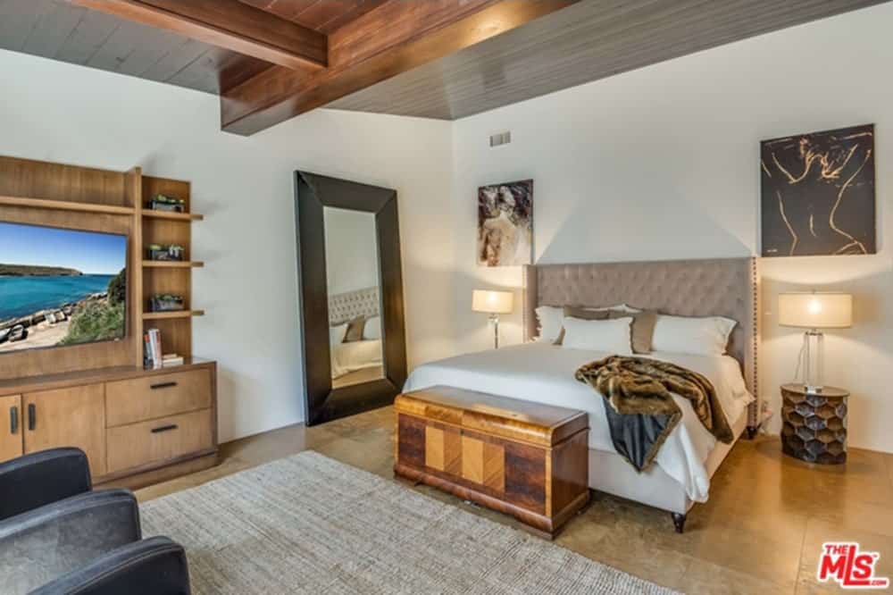 A black framed mirror faces the tufted wingback bed situated in between stylish nightstands topped with drum table lamps. It has lovely wall arts and a flat panel TV mounted on the wooden cabinet.