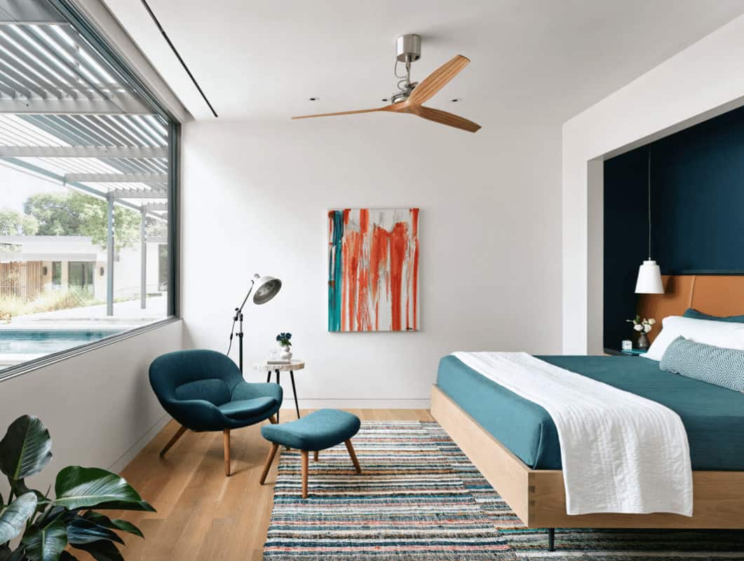 White primary bedroom with an abstract artwork and teal round back lounge chair that complements the cozy bed placed on the inset wall.