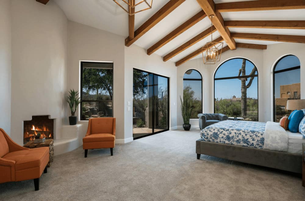 Spacious bedroom with orange tufted chairs and a gray velvet bed that matches the chesterfield chair by the arched windows. It is lighted by brass chandeliers that hung from the wood beam ceiling.