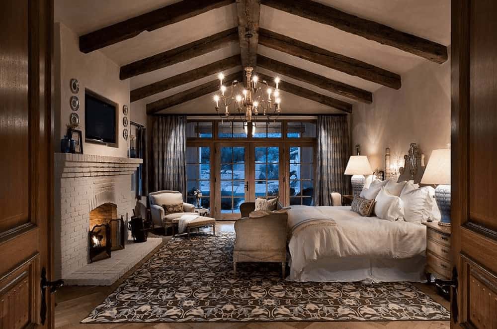 A candle chandelier illuminates this primary bedroom along with table lamps that sit on the wooden nightstands. It has a white skirted bed and a brick fireplace with a flat panel TV on top.
