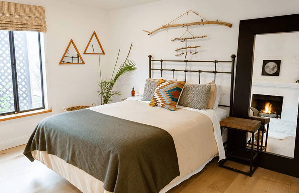 White primary bedroom decorated with triangular mirrors and wood wall arts that hung above the metal bed accented with an eye-catching throw pillow. It includes a moon artwork and a white fireplace reflected in the black framed mirror.