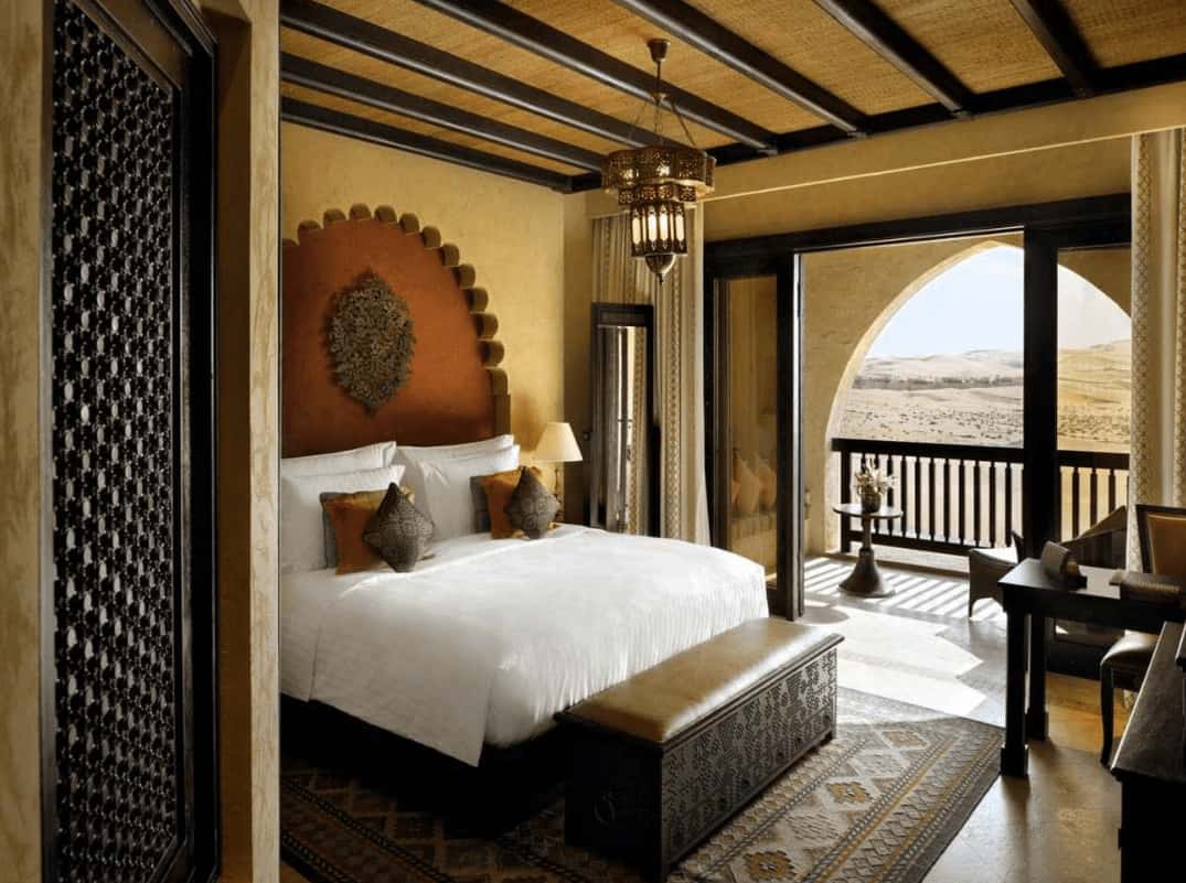 An intricate cushioned bench sits in front of the gorgeous bed lighted by a vintage chandelier that hung from the wood beam ceiling. It has classic area rug and glass sliders that open to the balcony.