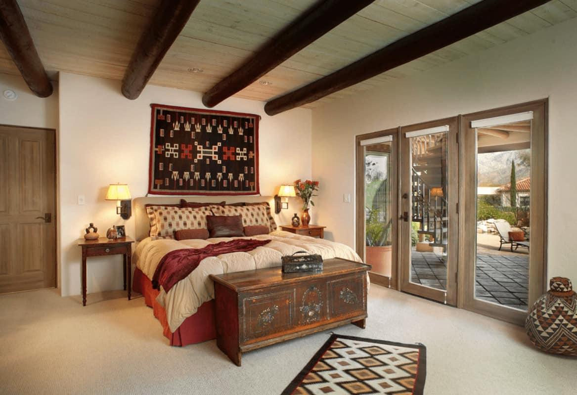 Southwestern bedroom showcases wood beam ceiling and carpet flooring topped by a patterned runner. It has a black tapestry and red skirted bed with a rustic console table on its end.