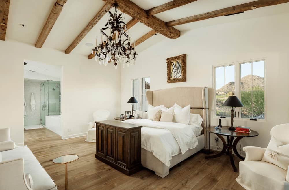 White primary bedroom with wide plank flooring and glazed windows framing a spectacular mountain view. It has comfy seats and a beige wingback bed lighted by a fabulous chandelier.