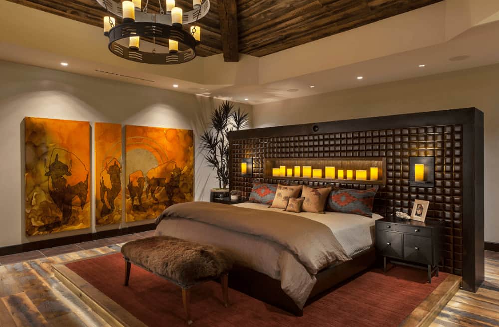 Warm bedroom decorated with multi-panel wall art and a two-tier candle chandelier that hung from the wood beam ceiling. It has a furry bench and a wooden bed with a gorgeous custom headboard.