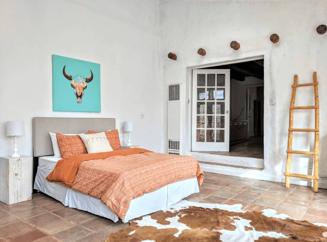 A bull head artwork adds a nice accent in this white bedroom with a gray skirted bed and cowhide rug that lays on the terracotta flooring.