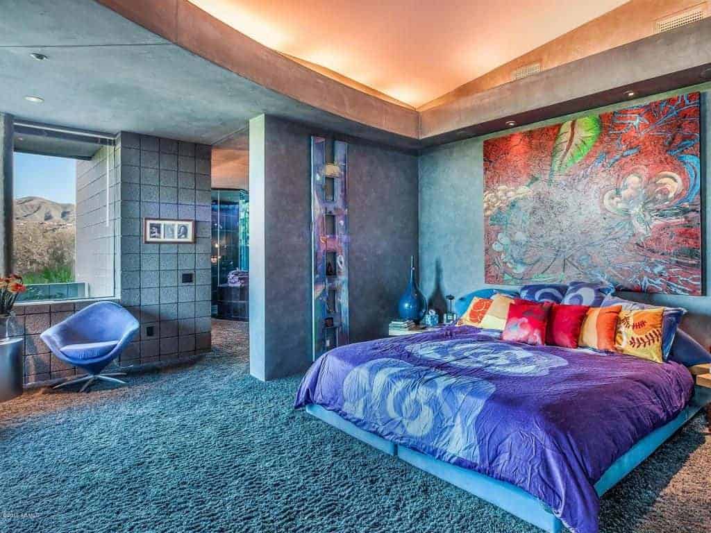 Marvelous bedroom with carpet flooring and a picture window framing an amazing mountain view. It has a round back chair and velvet bed with large artwork on top.