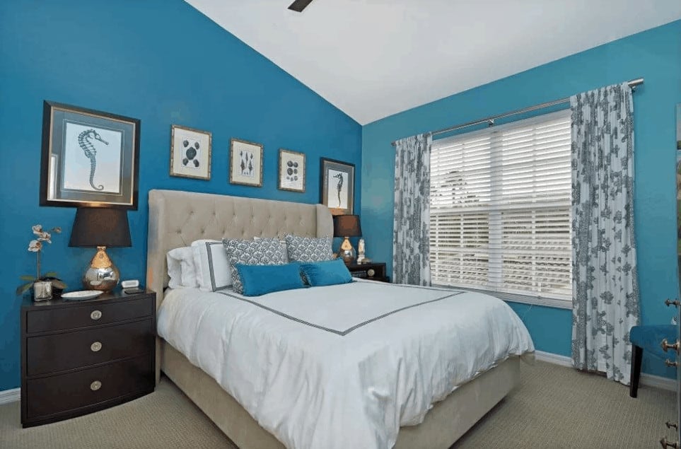 This primary bedroom features a tufted wingback bed in between wooden nightstands lined with framed wall arts. It has carpet flooring and glazed windows covered in floral draperies.