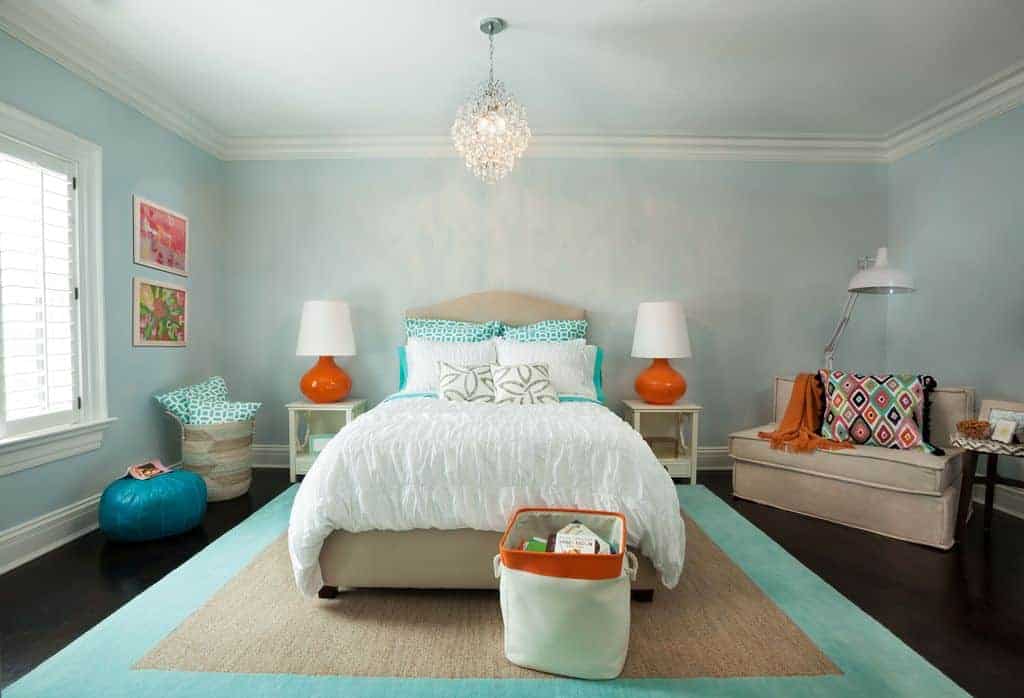 Muted blue bedroom with subtle orange accents from the table lamps, throw blanket and storage bin. It has a beige bed on a jute bordered rug that complements the upholstered seat topped with an eye-catching patterned pillow.
