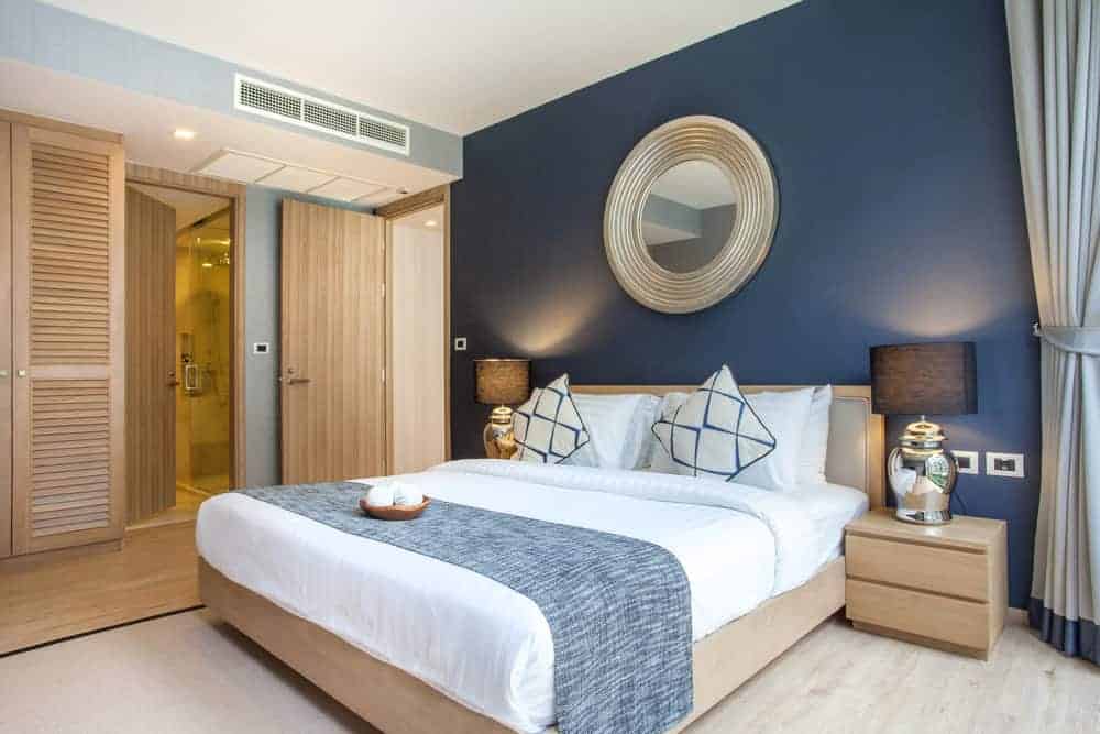 A wooden door opens to this blue primary bedroom showcasing a louvered wardrobe and comfy bed accented by a round mirror.