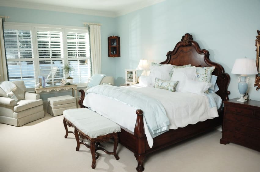 Lovely table lamps sit on the dark wood nightstands in this primary bedroom with gray skirted chairs and a carved wood bed with a cushioned bench on its end.