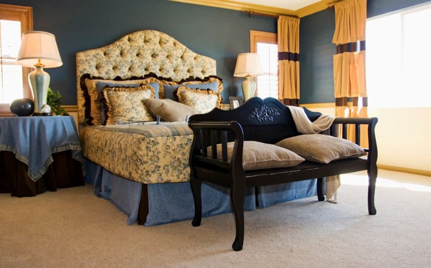 A black bench sits in front of the gorgeous tufted bed dressed in floral bedding. It is flanked by skirted nightstands and lovely table lamps.