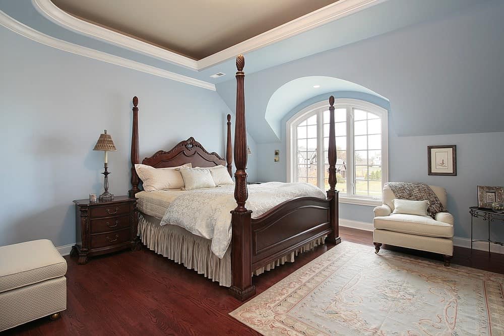 Charming primary bedroom with a tray ceiling and rich hardwood flooring topped by a light blue rug. It has beige seats and a four poster bed that complements the wooden nightstands.