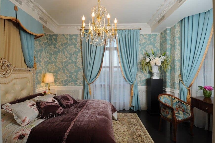 Elegant primary bedroom clad in blue floral wallpaper and white wainscoting. It has a cream tufted bed and a dark wood desk paired with a printed round back chair.