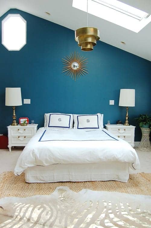 Gorgeous primary bedroom decorated with layered rugs and a sunburst mirror mounted above the white skirted bed. It has carpet flooring and a vaulted ceiling fitted with a skylight.