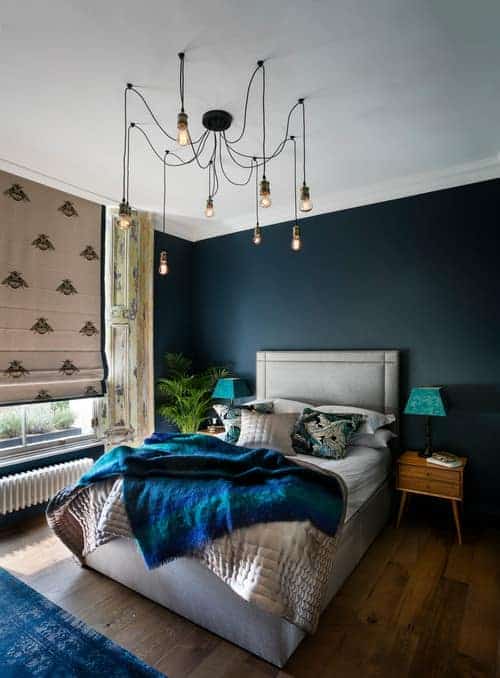 An industrial chandelier illuminates this blue bedroom featuring a gray upholstered bed and wooden nightstands topped with blue lampshades.