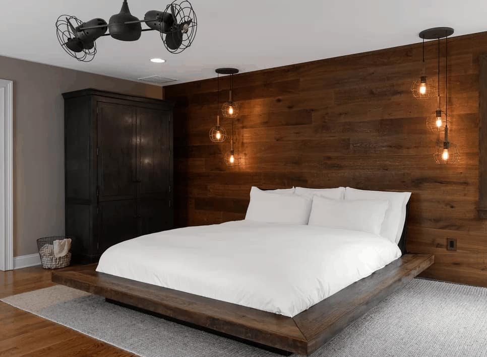 A black ceiling fan complements the wardrobe cabinet that faces the platform bed on a gray area rug with a custom wood plank headboard lighted by bulb pendants.