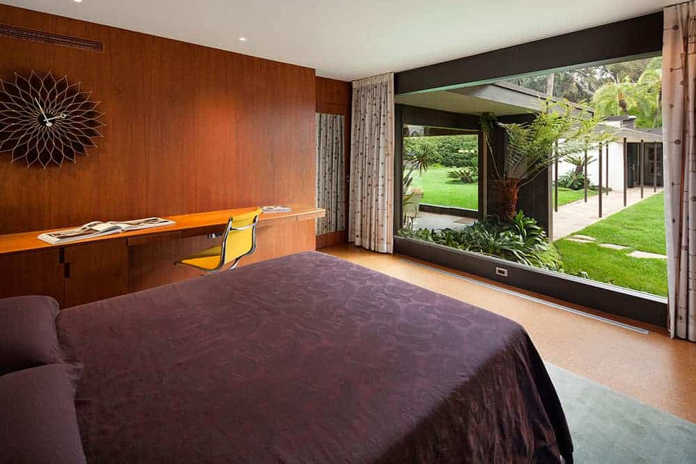 Brown primary bedroom with a comfy bed and a stylish wall clock that hung above the floating desk paired with a yellow swivel chair. It has wood paneled walls and a panoramic window overlooking the lush green garden.