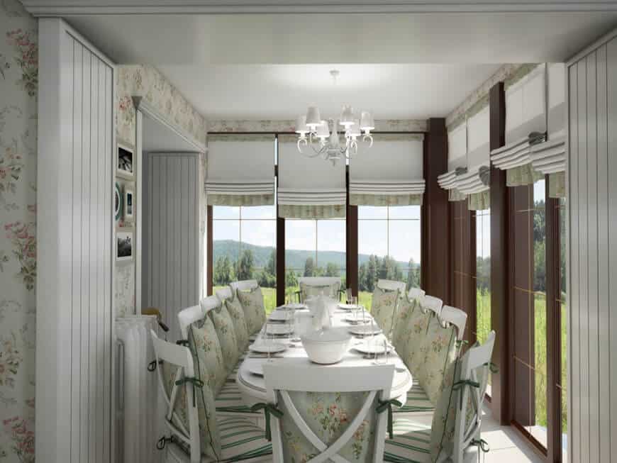 This country-style dining room has white cross-back chairs with light green floral cushions that match with the wallpaper and the shades of the surrounding tall windows that showcase a lush green landscape outside.
