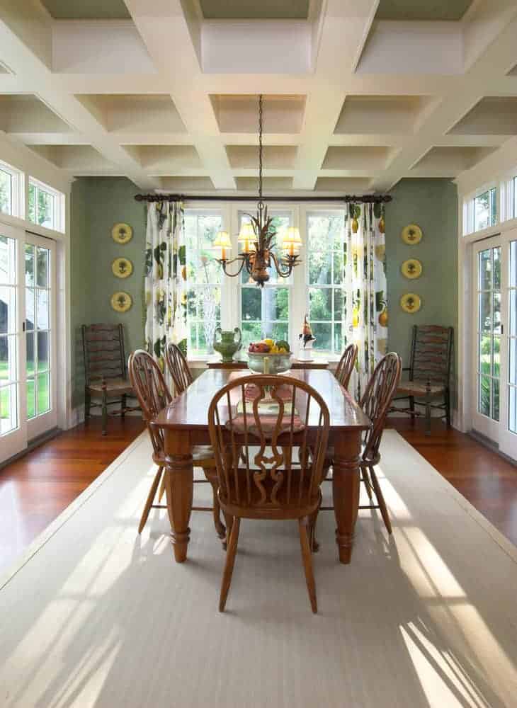 The traditional wooden dining set is separated from the from by a light gray area rug. This hue of the rug is closely related to the coffered ceiling that has a simple chandelier hanging over the table that is complemented by the green walls.