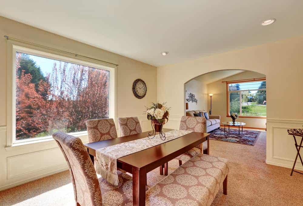 The charming chic floral cushions of the Parson's chairs and bench blends with the salmon pink carpeted flooring. This hue is complemented by the dark wooden dining table and the abundant natural lights coming in from the wide glass window.