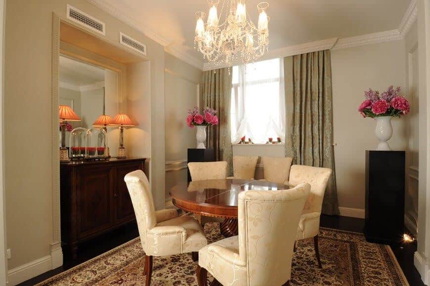 This dining room has a classy chic demeanor to its intimate setup of small wooden dining table and its four beige cushioned chairs that has subtle floral patterns to match with the chandelier and patterned area rug that covers most of the dark hardwood flooring.