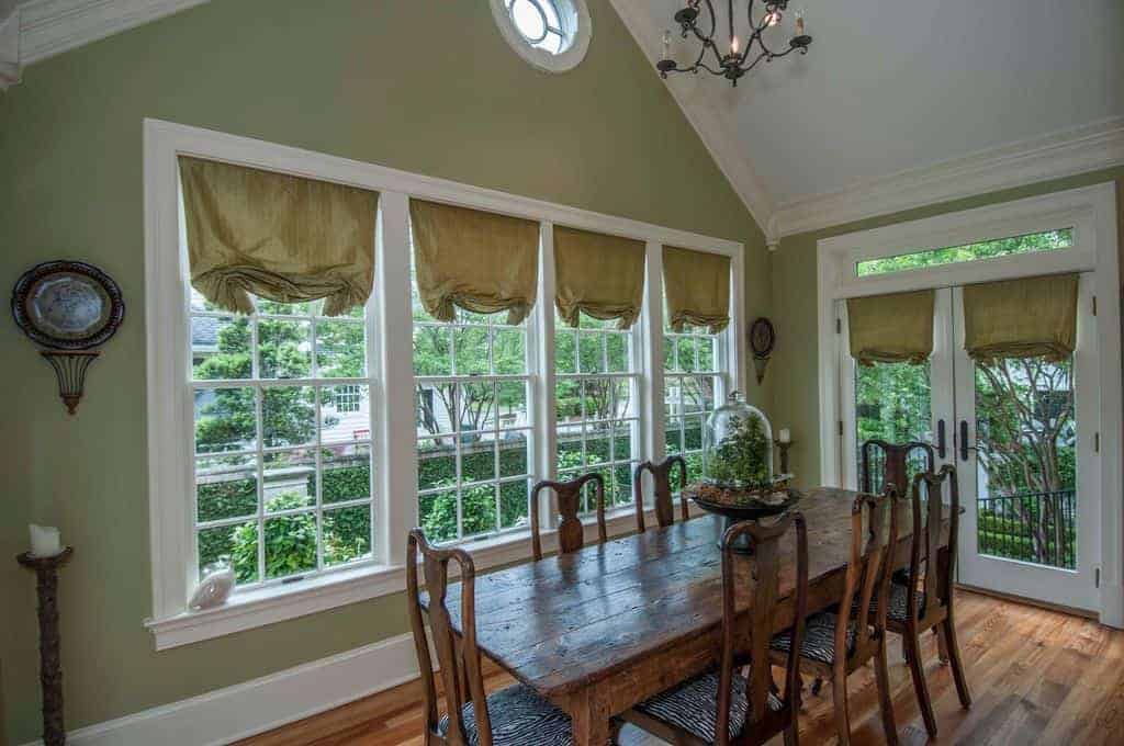 This simple country-style dining room has a white cathedral ceiling that bears a wrought iron chandelier over the wooden dining table and its wooden chairs that match with the hardwood flooring and complemented by the green walls and its glass windows.