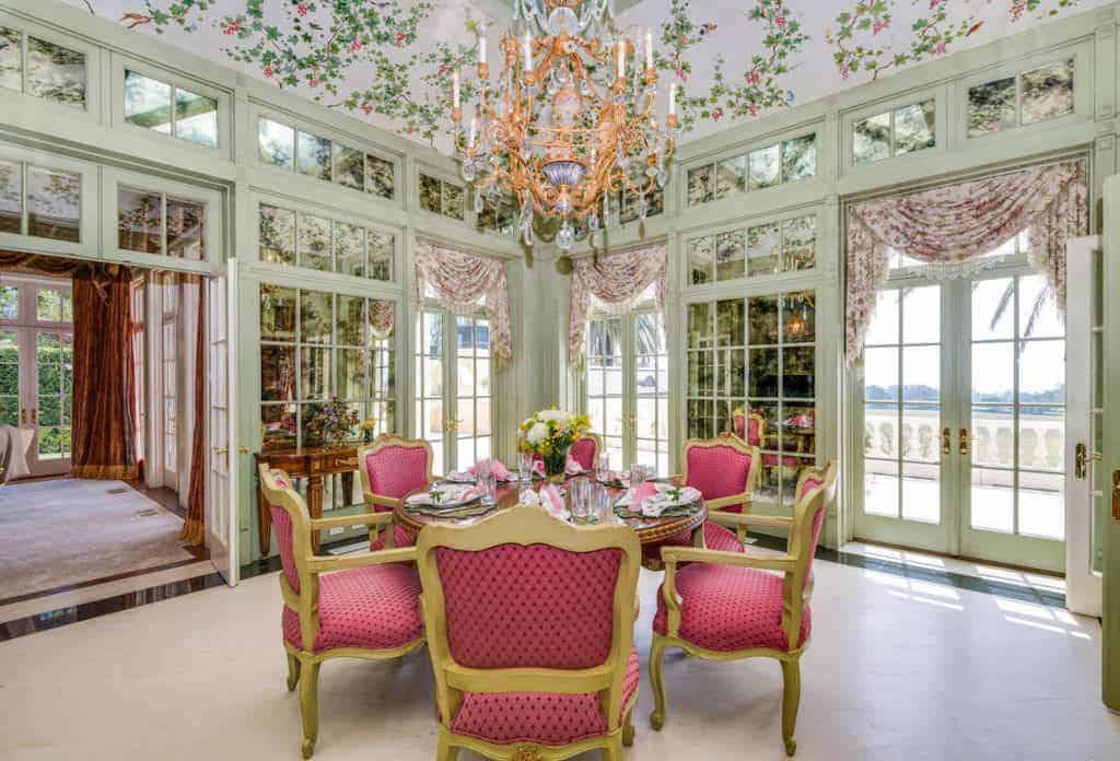 The elegant dining chairs surrounding the round wooden table has golden frames and pink patterned cushions that stand out against the light gray carpeted flooring. This simple flooring is contrasted by the floral ceiling with a majestic chandelier.