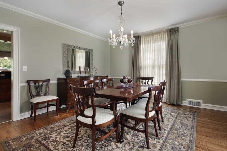The light green walls makes the dark wooden dining set stand out as well as its colorful patterned area rug that is illuminated by the curtained window by the head of the table. The window is augmented by the crystal chandelier.