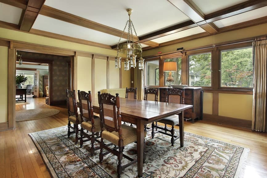 The charming patterned area rug complements the rectangular wooden table and its dining chairs that has woven wicker backs and beige seats. This is paired with a golden chandelier hanging from the white coffered ceiling.