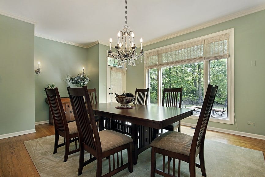 The dark wooden legs of the rectangular dining table matches with the design of the slat backs of the dining chairs. This setup is contrasted by the white ceiling that hangs a wrought iron chandelier matching with the wall mounted lamps of the green walls.