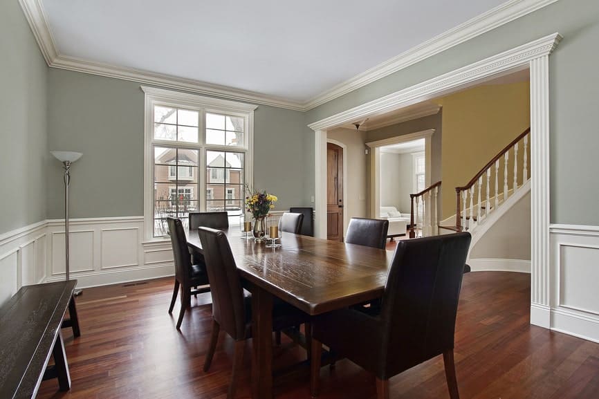 The light gray walls of this dining room is complemented by the white wainscoting that contrasts the dark hardwood flooring and the dark wooden bench pressed against the wall beside the wooden dining set that has black leather cushions on its chairs.