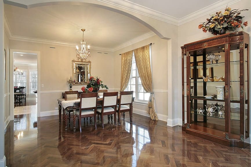 The sleek hardwood flooring has a herring bone pattern that brings a complex and shiny background for the elegant dining set with a dark brown hue and beige cushions. This pairs well with the dining room cabinet that is mostly made of glass.