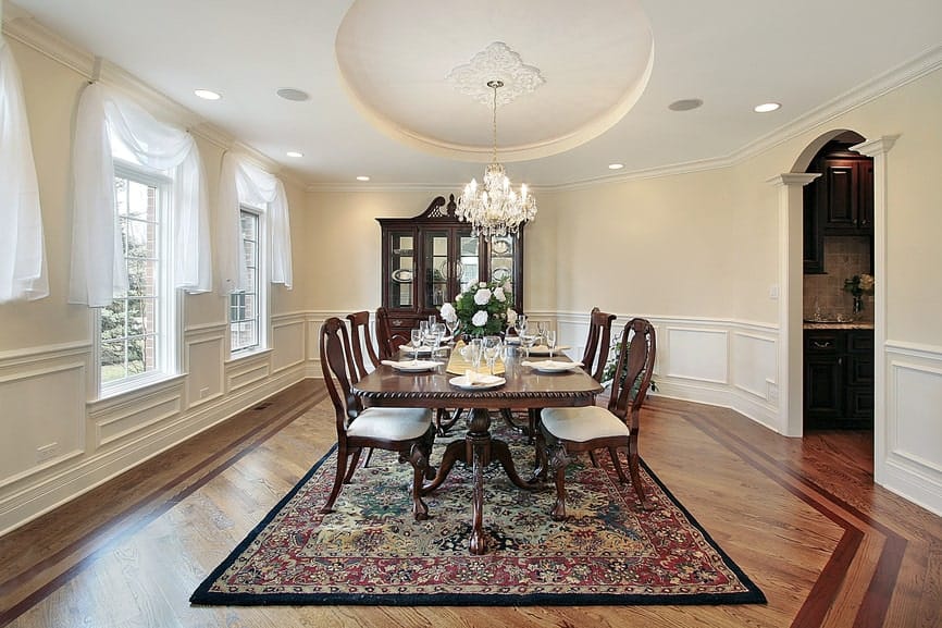 The spacious hardwood flooring is mostly covered with a colorful area rug that complements the dark wooden elements of the elegant dining table and its wooden chairs contrasted by the white chandelier from the circular tray of the ceiling.