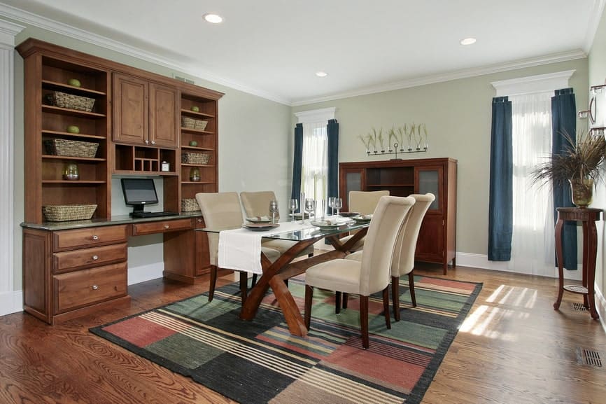 This country-style dining room has a glass-top dining table over a colorful area rug that tops the hardwood flooring. This blends with the table frame and the dining room cabinets that stand out against the light green walls.