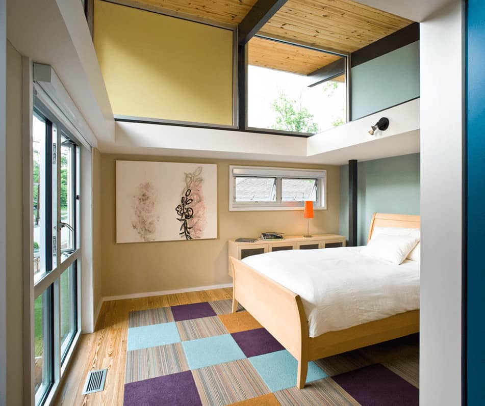 Contemporary bedroom with multi-colored walls and glass paneled windows allowing natural light in. It has lovely artwork and a light wood bed that sits on a checkered area rug.