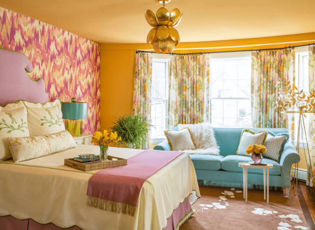 Charming bedroom showcases a pink skirted bed and blue sectional sofa filled with fluffy pillows. It is illuminated by a gold chandelier along with natural light that flows in through the bay window. pink skirted bed and blue sectional sofa filled with fluffy pillows. It is illuminated by a gold chandelier along with natural light that flows in through the bay window.