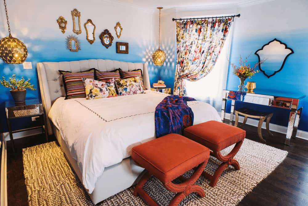 A pair of brass pendant lights hang over the mirrored nightstands in this fabulous bedroom with dark hardwood flooring and blue ombre walls mounted with various styled mirrors.