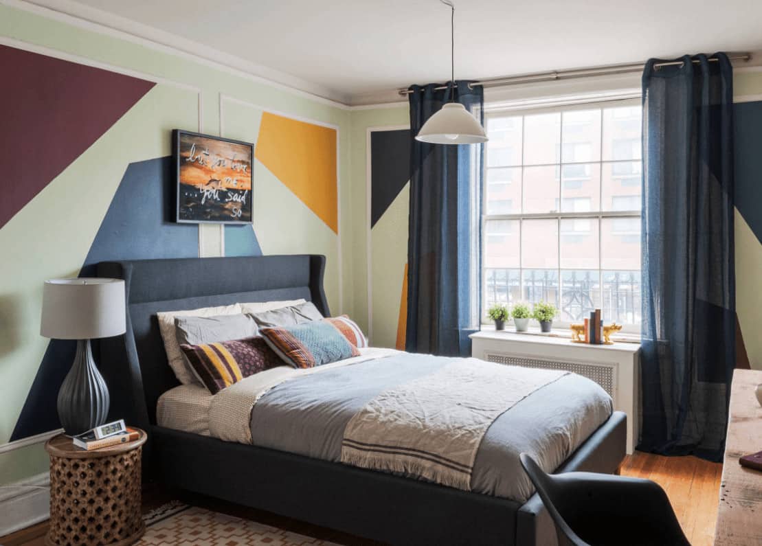 A bachelor pad with a white framed window and wainscoted walls painted in geometric design. It has a white dome pendant and navy blue bed paired with a round nightstand.