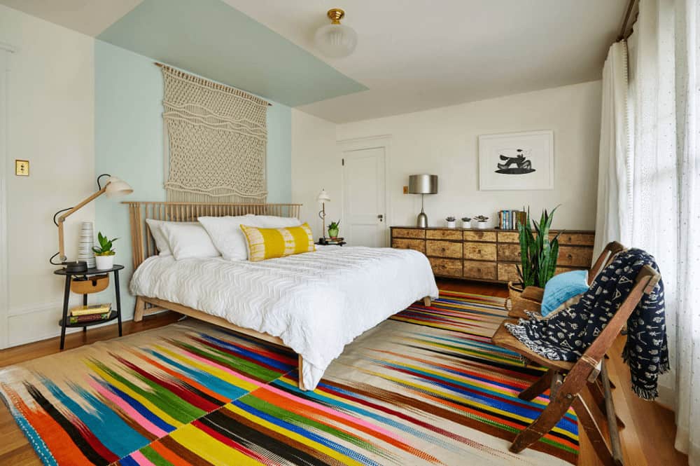 An eye-catching multi-colored rug adds a striking accent in this primary bedroom showcasing a wooden bed and long distressed dresser topped with a chrome table lamp.