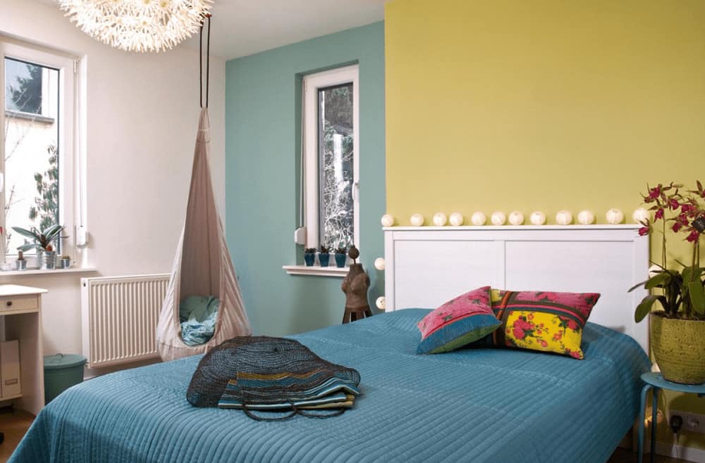 A lovely potted plant sits next to the white bed dressed in teal bedding in this multi-colored bedroom boasting a fabric hanging chair and glazed windows that invite natural light in.