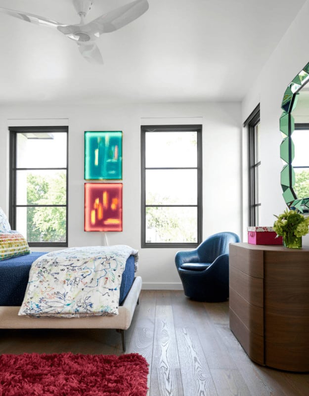 A white ceiling fan hangs over the upholstered bed facing the wooden dresser and stylish mirror. It is accompanied by a blue round back chair and a red shaggy rug.